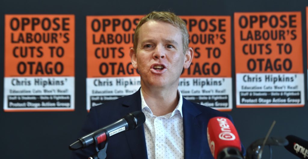 Hipkins in front of posters that read 'Oppose Labour's Cuts to Otago'